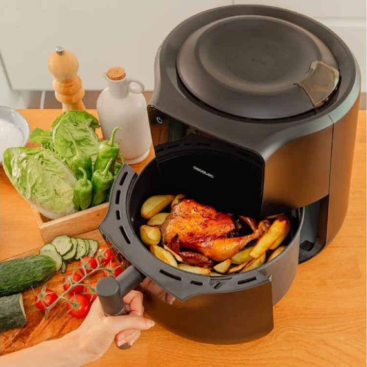 Black Cecotec air fryer with open basket containing roasted chicken and fries, on wooden table with vegetables.