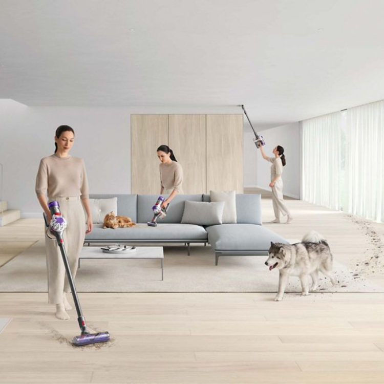 Dyson V8 silver and nickel vertical vacuum cleaner in use by a woman in a living room.