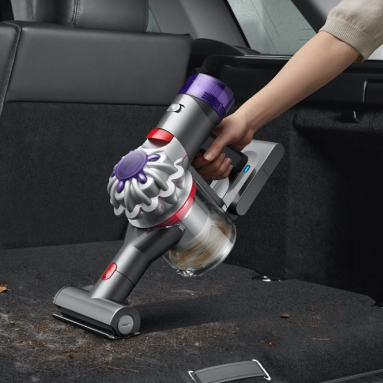 Dyson V8 handheld vacuum cleaner, silver and purple, being used to clean car interior.