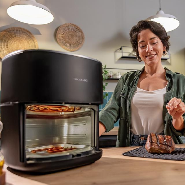 Black Cecotec air fryer with glass door, woman smiling beside it, grilled meat on tray.