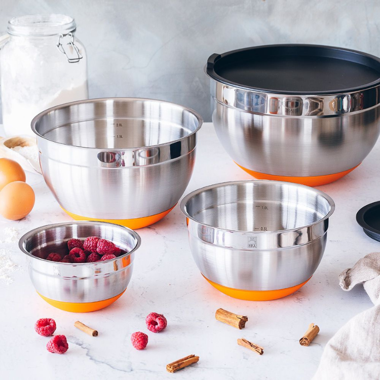 Stainless steel bowl set with orange bases, one with raspberries, on a marble countertop with eggs, flour, and spices.