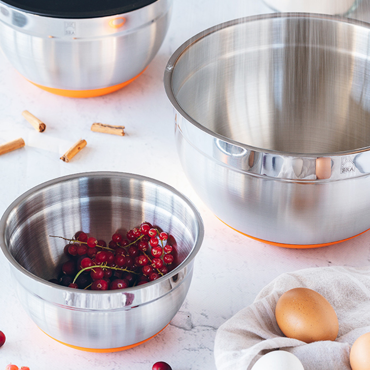Stainless steel bowl set with orange base, one containing red currants, on a white marble countertop with eggs and cinnamon rolls.
