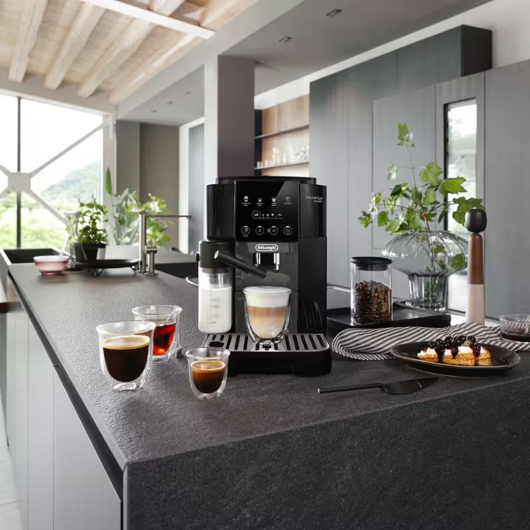 Black Delonghi Magnifica Start LatteCrema coffee machine on kitchen counter with four cups of coffee and pastries.
