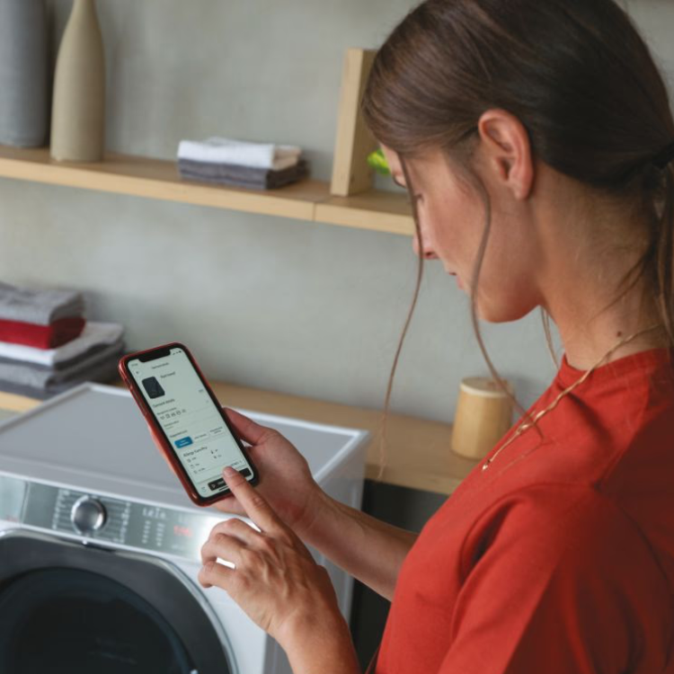 Woman in red shirt using smartphone next to white Hoover washing machine.