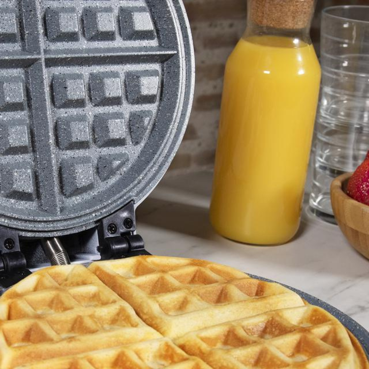 Cecotec Fun Gofrestone 3-in-1 waffle maker with waffles, orange juice, and strawberries on a kitchen counter.