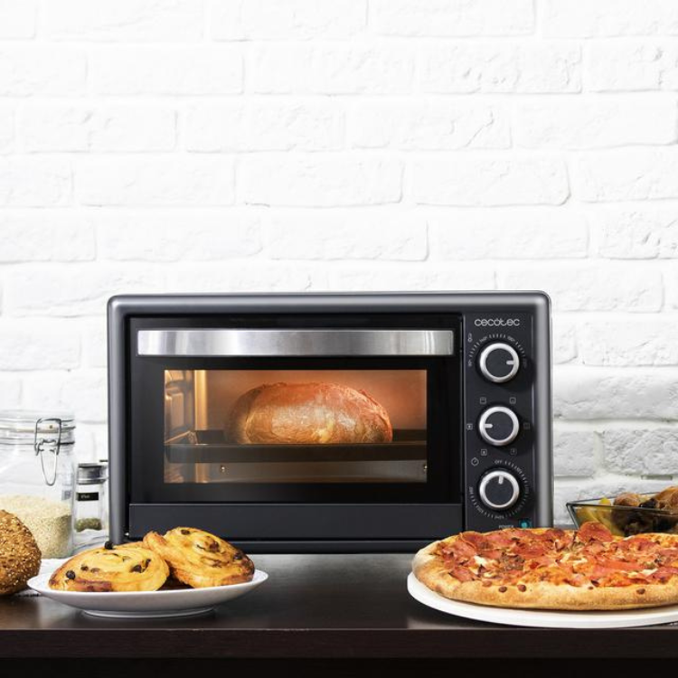 Black Cecotec mini oven with silver accents, 3 dials, and glass door, surrounded by pizza, bread rolls, and bread on a table.
