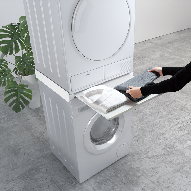 White stacked washer and dryer with foldable tray, hands placing clothes on it. Napofix ML-3 vertical mounting base.