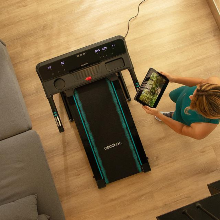 Black Cecotec DrumFit WayHome 1000 Runner Sprint treadmill with teal accents and digital display, woman holding tablet beside it.
